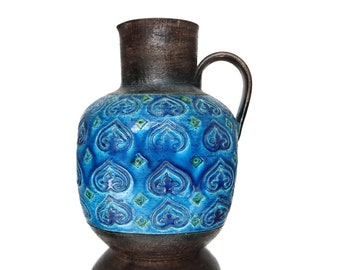 May include: A blue and brown ceramic pitcher with a decorative pattern of stylized hearts. The pitcher has a wide base and a narrow neck with a handle.