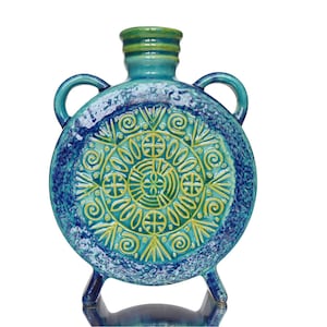 May include: A circular ceramic flask featuring a vibrant teal and blue glaze with a textured finish. The front displays a raised, intricate yellow geometric pattern of concentric circles and radiating lines. It has two side handles and a short neck.