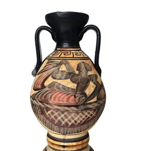 Könnte beinhalten: Eine schwarze und braune Keramikvase mit einem bemalten Design einer Frau in einem fließenden Gewand. Die Vase hat zwei Henkel und einen runden Körper.