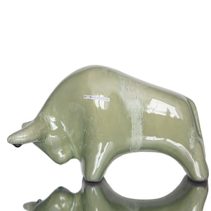 Puede incluir: Una figurita de toro de cerámica verde con un acabado brillante. La figurita está de pie sobre una superficie blanca y tiene una etiqueta que dice "GTO KERAMIK".