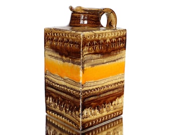 May include: A square ceramic pitcher with a brown, orange, and beige glaze. The pitcher has a geometric pattern and a wide mouth.