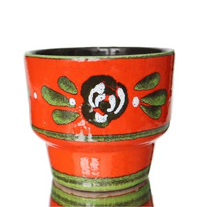 May include: A bright orange ceramic plant pot with a black interior and a floral design. The flower is white with a black centre, surrounded by green leaf-shaped accents. The pot has a glossy finish.