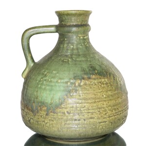 Könnte beinhalten: Ein grüner Keramikkrug mit Henkel. Der Krug hat einen runden Körper und einen schmalen Hals. Die Glasur hat ein gesprenkeltes Aussehen mit Grüntönen und Braun. Es ist wahrscheinlich ein dekorativer oder funktionaler Gegenstand für das Zuhause.