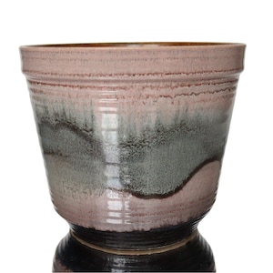 Może przedstawiać: Ceramiczna donica z szerokim, teksturowanym brzegiem i rozszerzanym korpusem. Donica ma gradient kolorów, w tym różowy, szary i brązowy, z ciemnobrązową podstawą. Mierzy około 25 cm wysokości.