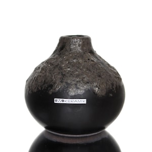 Könnte beinhalten: Eine schwarze Keramikvase mit einem strukturierten, dunkelgrauen oberen Teil und einem glatten, abgerundeten Boden. Die Vase hat einen schmalen Hals und ein Etikett mit der Aufschrift "OTTO KERAMIK". Die Vase wird vor weißem Hintergrund gezeigt.