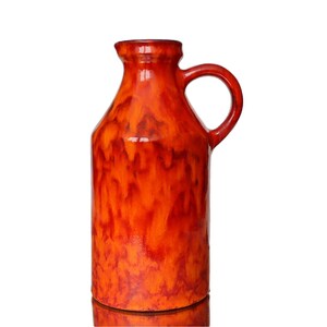 Op de afbeelding: Een hoge, oranje keramische vaas met een handvat. De vaas heeft een glanzende afwerking met een gevlekt patroon van donkerrode en oranje tinten. De vaas is ongeveer 30 cm hoog.