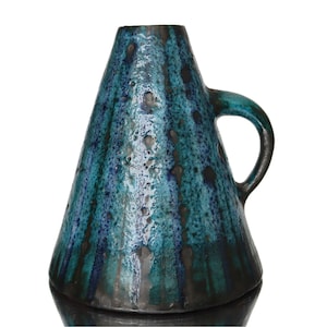 May include: A teal and dark blue ceramic vase with a handle. The vase has a unique shape, wider at the base and tapering upwards. The glaze has a textured, speckled appearance with dark streaks.