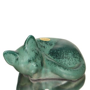 Op de afbeelding: Een keramieken beeldje van een slapende kat, opgerold. De kat heeft een gevlekte groene kleur met donkerdere groene accenten. Het beeldje heeft een glanzende afwerking en rust op een reflecterend oppervlak.