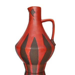 Puede incluir: Jarra de cerámica roja con asa y base ancha. La jarra presenta rayas verticales de color marrón oscuro sobre un fondo rojo. La jarra tiene una forma única y un área ligeramente astillada cerca de la parte superior.