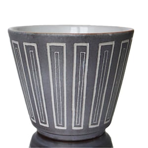 Könnte beinhalten: Eine graue Keramikschale mit weißem geometrischem Muster. Die Schale hat eine ausgestellte Form und einen weißen Innenraum. Die Außenseite weist vertikale rechteckige Designs auf. Die Schale steht auf einem dunklen Sockel.