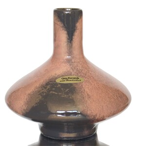 Könnte beinhalten: Eine braune und schwarze Keramikvase mit breiter Basis und schmalem Hals. Die Vase hat eine gesprenkelte Glasur und ein Etikett mit der Aufschrift "Otto Keramik echt Handarbeit".