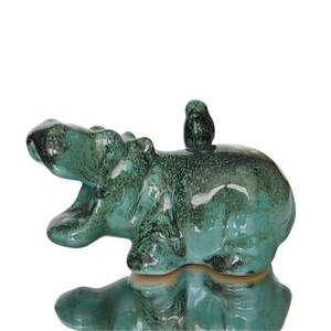 Könnte beinhalten: Eine Nilpferd-Figur aus Keramik mit einer glänzenden, blaugrün und dunkelgrün gesprenkelten Glasur. Das Nilpferd ist mit weit geöffnetem Maul dargestellt, und ein kleiner Vogel sitzt auf seinem Rücken. Die Oberfläche hat ein glattes Finish.