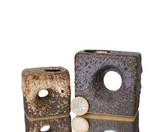 May include: Two square ceramic vases with a hole in the center. The vase on the left is brown and white with a rough texture. The vase on the right is purple and brown with a rough texture. A gold coin is in front of the vases.