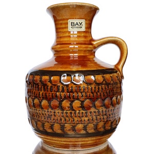May include: A brown ceramic vase with a geometric pattern of circles and lines. The vase has a handle and a label that reads "BAY Keramik".