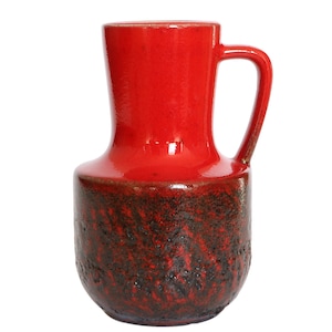 Può includere: Un vaso in ceramica rosso con una glassa marrone e un manico. Il vaso ha un corpo arrotondato e un collo stretto.