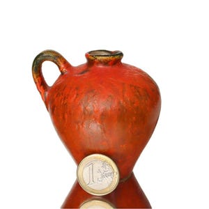 Può includere: Un piccolo brocca in ceramica arancione con una superficie strutturata. La brocca è grande circa come una moneta da un euro.