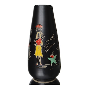 May include: Black ceramic vase with a decorative relief of a woman carrying a basket on her head. The woman is wearing a red top and yellow skirt. The vase has a flared base and a narrow neck. The artwork is in a folk art style.