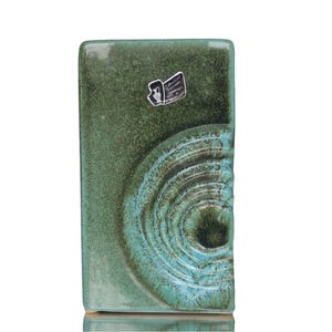 Puede incluir: Un jarrón de cerámica verde con un patrón de espiral en la parte delantera. El jarrón tiene una forma rectangular y una superficie texturizada.