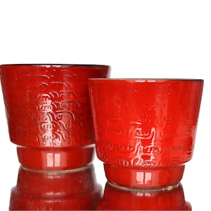 Peut inclure: Deux cache-pots en céramique rouge vif avec une finition brillante et un motif en relief répétitif représentant des visages stylisés. Les pots cylindriques présentent une base légèrement effilée et un large sommet ouvert.