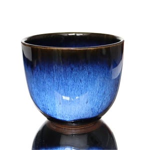 May include: A rounded ceramic cup featuring a glossy glaze that transitions from a deep, dark brown at the rim to a vibrant, mottled cobalt blue in the centre. The vessel has a smooth, curved silhouette and a wide opening, showcasing a reactive finish.