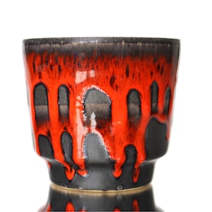 Könnte beinhalten: Eine Keramikvase mit auffälligem Design. Der obere und untere Bereich sind schwarz, während der mittlere Bereich eine leuchtend rote Glasur aufweist, die heruntertropft und einen einzigartigen visuellen Effekt erzeugt. Die Vase hat eine glänzende Oberfläche und eine weite Öffnung.