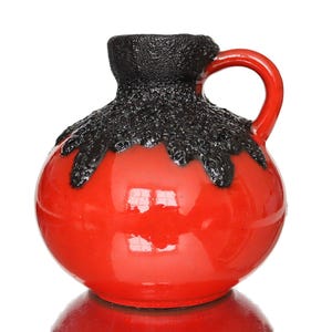 May include: A round ceramic pitcher featuring a glossy, vibrant red finish on the lower body and a contrasting, textured, matte black glaze dripping from the rim down to the shoulder. The vessel includes a single curved handle on the right side.