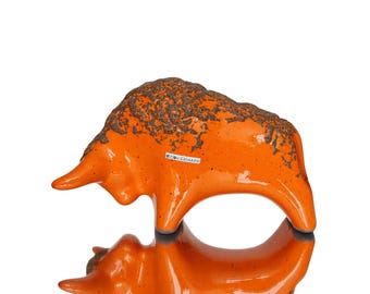 Pode incluir: Uma estatueta de touro em cerâmica laranja brilhante com um acabamento texturizado castanho escuro salpicado nas costas. O touro é representado numa posição de ataque com a cabeça baixa. Uma pequena etiqueta branca diz OMO KERAMIK.