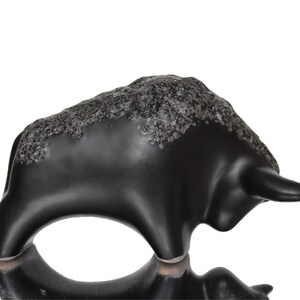May include: Black ceramic bull figurine with a textured surface. The bull is standing on its four legs with its head turned to the side.