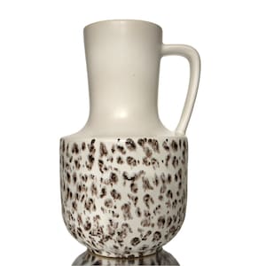 May include: Cream-colored ceramic pitcher with a handle. The lower portion of the pitcher features a speckled pattern in shades of brown. The upper portion and handle are a solid cream color. The pitcher has a rounded base and a slightly flared top.