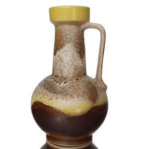 May include: A vintage ceramic pitcher with a handle. The pitcher features a bulbous base and a cylindrical neck, with a yellow rim and handle. The body is a mix of brown, tan, and yellow hues, creating a textured, layered effect.