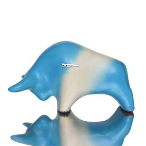 May include: A ceramic bull figurine in a charging pose, featuring a smooth gradient finish that transitions from bright blue at the head and rear to a creamy white midsection. A small rectangular label on the side reads OKERAMIK.
