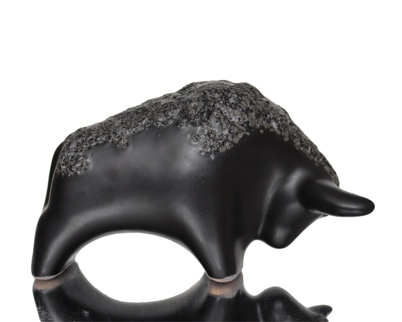 May include: Black ceramic bull figurine with a textured surface. The bull is standing on its four legs with its head turned to the side.
