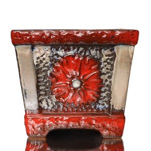 May include: A red and white ceramic planter with a raised floral design. The planter has a square shape with rounded edges and a textured surface.
