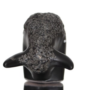 May include: Black ceramic whale sculpture with a textured surface. The whale is facing forward with its mouth closed and its tail fin raised.