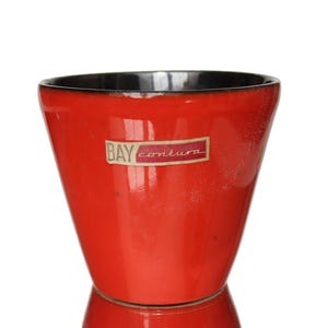 Peut inclure: Un cache-pot en céramique rouge vif avec une finition brillante et un intérieur sombre. L'extérieur présente une étiquette rectangulaire avec le texte BAY contura imprimé en blanc et noir. Le récipient a un bord évasé et un corps cylindrique.