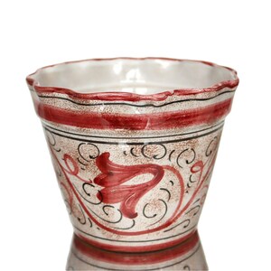 May include: A ceramic flower pot with a scalloped rim. The pot features a cream-colored base with red and black painted floral designs and bands. The interior is white, and the pot has a slightly flared shape.