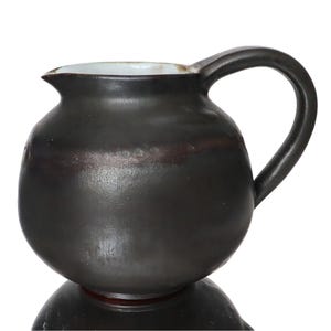 Può includere: Una brocca in ceramica grigio scuro con un corpo arrotondato e un manico curvo. La brocca ha un beccuccio e un bordo interno bianco. La base della brocca si riflette sulla superficie sottostante.