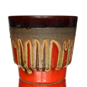 Può includere: Un vaso per piante in ceramica dalla forma cilindrica larga con un bordo marrone scuro. Il corpo presenta uno smalto testurizzato tipo lava con gocciolature verticali color marrone e beige che scendono su una base arancione vivace.