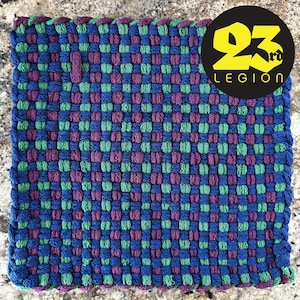 May include: A woven square mat with a checkered pattern in shades of blue, green, and purple. The mat is made of thick yarn and has a fringed edge. The mat is sitting on a light brown surface.