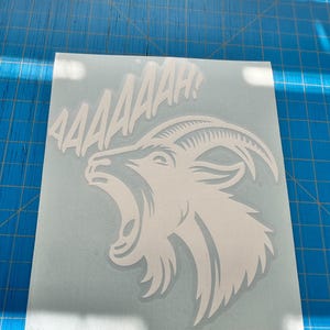 May include: White vinyl decal of a screaming goat with the word "AAAAAAH!" above it. The goat has an open mouth and curved horns. The decal is on a light blue background, ideal for applying to various surfaces.
