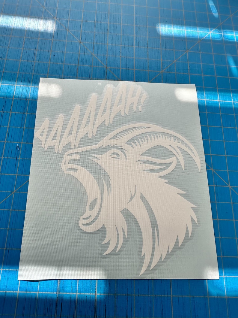 May include: White vinyl decal of a screaming goat with the word "AAAAAAH!" above it. The goat has an open mouth and curved horns. The decal is on a light blue background, ideal for applying to various surfaces.