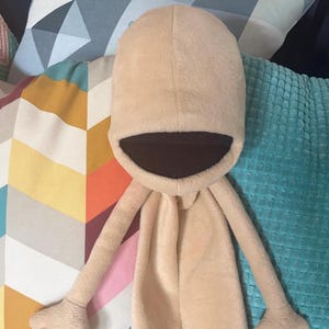 May include: A beige plush puppet with a large, oval head and a wide, dark brown mouth. The puppet has long, slender arms with beige hands. The puppet is designed to be held and manipulated, likely for entertainment or educational purposes.