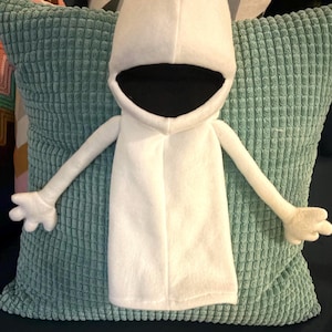 May include: A white felt puppet with a hooded head and a large black mouth. The puppet has long arms with three-fingered hands. It is resting on a textured, light teal pillow.