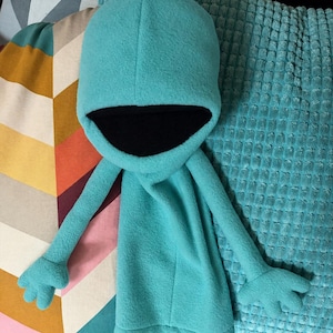 Puede incluir: Una prenda con capucha de forro polar turquesa que presenta mangas largas con extremos en forma de mano de cuatro dedos y un forro interior negro profundo en la capucha. La pieza está confeccionada con una tela suave y lisa de color sólido.
