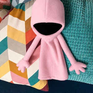 Op de afbeelding: Een zachte roze knuffel in de vorm van een cape met capuchon, lange armen en handen met vier vingers. De capuchon is gevoerd met een effen zwarte stof, wat een diepe, donkere opening creëert die contrasteert met het lichtroze fleecemateriaal.
