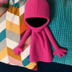 Puede incluir: Un peluche de forro polar rosa brillante que presenta una capucha redondeada con un interior negro profundo, brazos largos y delgados, y manos de cuatro dedos. La construcción de tela suave crea una textura lisa y un diseño de cuerpo liso.