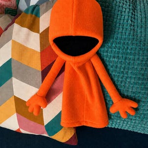 Puede incluir: Un peluche de color naranja brillante que presenta una capucha grande con un interior negro profundo, brazos largos y delgados, y manos de cuatro dedos. La figura tiene una textura simple y suave similar al forro polar y cabeza redondeada.