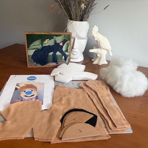 May include: A craft kit for a puppet featuring tan felt fabric pieces, black felt accents, white foam padding, a bag of white polyester fiberfill stuffing, and an instructional booklet with a photo of a finished puppet with a blue nose.