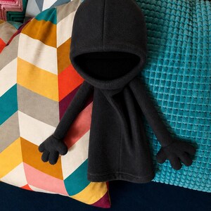 Könnte beinhalten: Ein schwarzes Plüschtier aus Fleece mit einer abgerundeten Kapuze, einem langen Körper und zwei ausgestreckten Armen mit vierfingrigen Händen. Der weiche, dunkle Stoff bedeckt die gesamte Figur, die eine glatte Textur und eine schlichte Form aufweist.