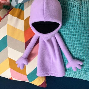Puede incluir: Un peluche hecho de tela polar de color morado claro con un diseño de capa con capucha, brazos largos, manos de cuatro dedos y un forro interior negro profundo dentro de la capucha, creando una figura de personaje simple, suave y redondeada.